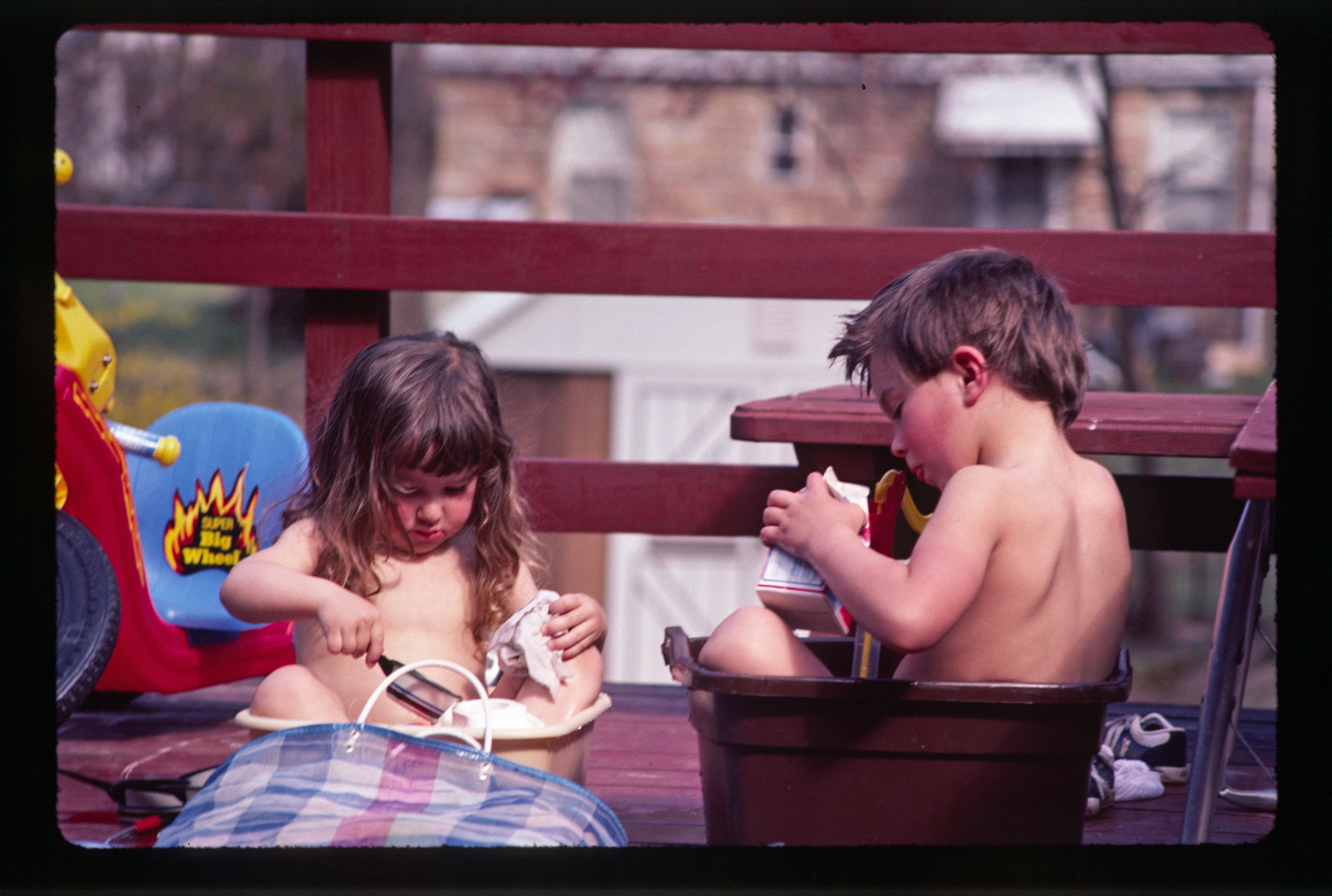 Young girl and boy on deck, each sitting in a small tub of water, they are intent on playing.